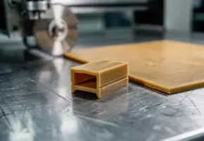 Precisiегesneden rubber- en kunststofonderdeel naast onbewerkte grondstof op stalen werkbank, met roterende snijschijf op achtergrond.