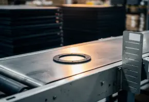 Rubberpakking op industriële transportband, omgeven door gestapelde materiaalbatches en een Kanban-kaarthouder op de machineframe.