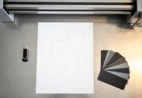 Technische tekening op wit papier naast USB-stick en materiaalstalen van rubber en plastic, met lasercutmachine op achtergrond.