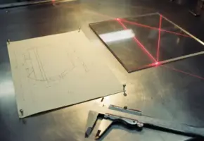 Technische tekening op rasterpapier naast een acrylplaat met rode laserbundel en industriële schuifmaat op stalen werktafel.