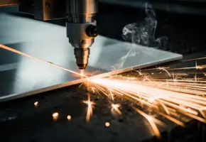 Lasersnijkop snijdt door gepolijst aluminiumplaat, oranje vonken spatten op donkere werkplaatsvloer, dunne rook kringelt omhoog.