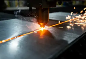 Lasersnijder snijdt door gegalvaniseerd staalplaat met oranje gloeiend snijpunt en opstijgende rook in industriële werkplaats.