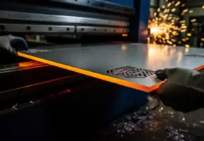 Lasergestanst stalen paneel met gloeiende randen in een metaalpers, lasspatten op donkere industriële werkvloer op de achtergrond.